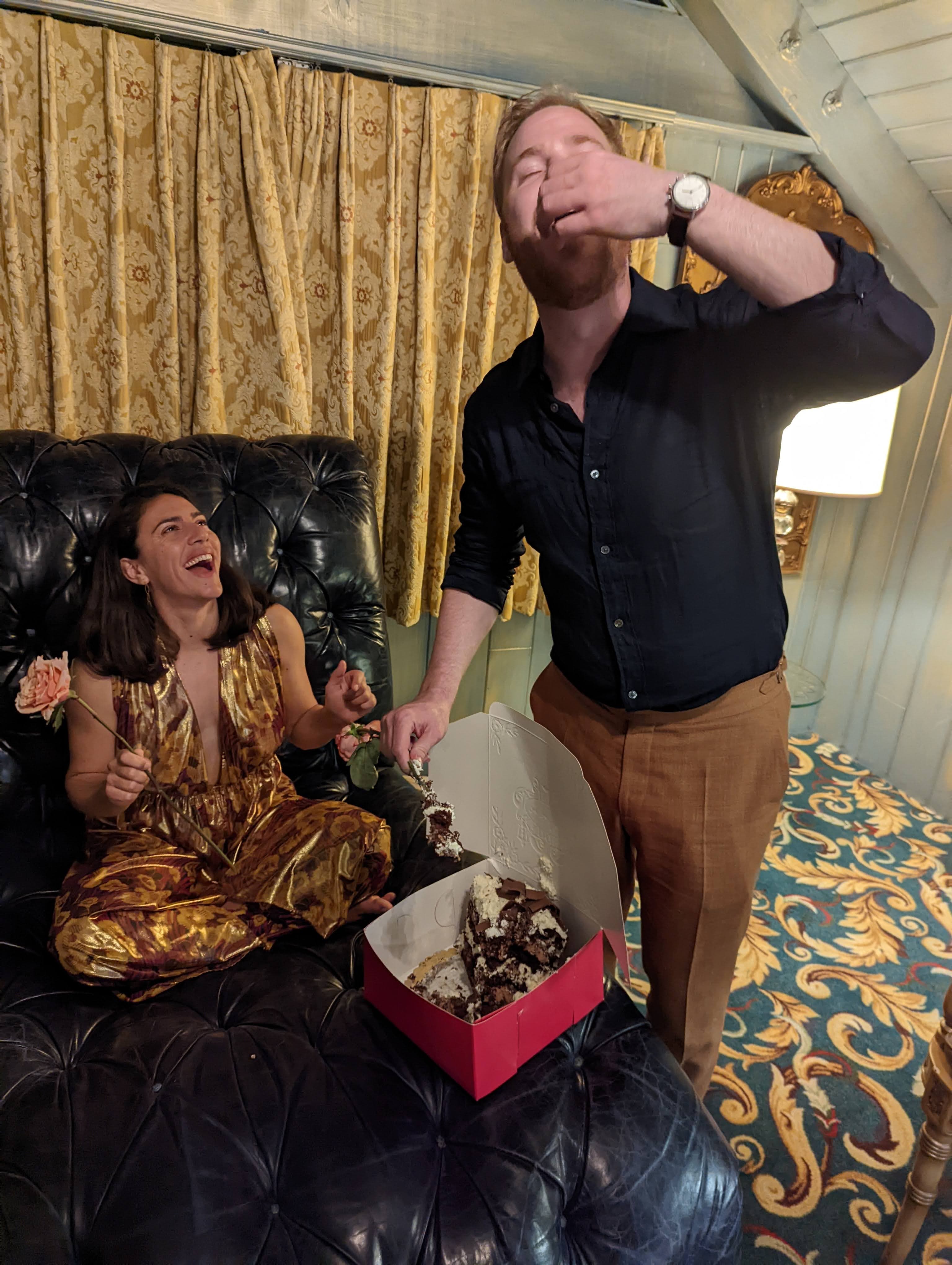 Ariana is sitting on a chair in a room at the Madonna Inn watching Erik eat a bite of cake